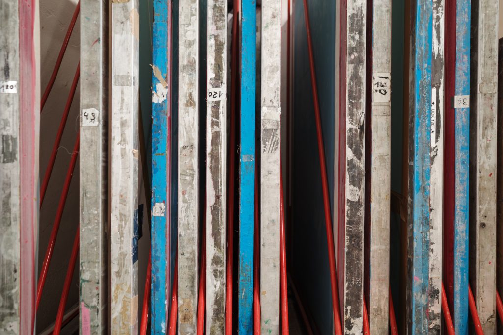 A close-up side view of used screen printing frames stored vertically in a red metal rack within an art studio. The aluminum and wooden frames are stained with dried blue and pink ink.