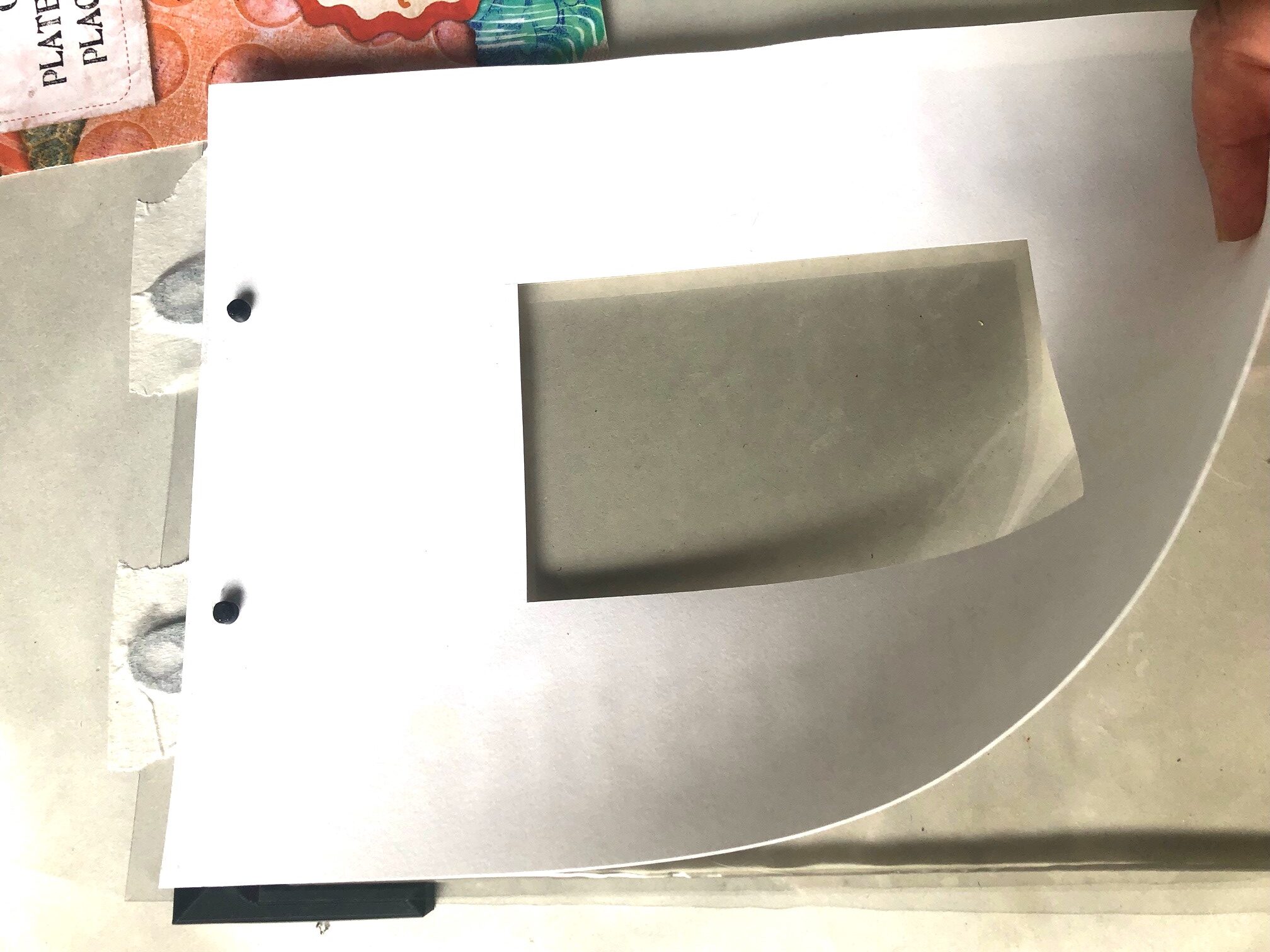 Black gel plate registration board, taped to work surface, shown with Gelli plate and hole punched white paper stencil